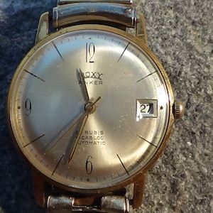 May include: A gold-toned wristwatch with a white face and black hands. The watch has a date window at the 3 o'clock position and the text "ROXY ANKER 21 RUBIS CABLOC AUTOMATIC" on the face.