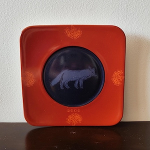 May include: A square plate with a deep red border and a dark blue circular center. The center features a light blue fox illustration. The red border is decorated with four orange floral motifs and the year 2000 printed at the bottom center.