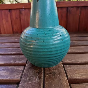 Puede incluir: Un jarrón de cerámica turquesa con base bulbosa y cuello estrecho. El jarrón tiene estrías horizontales alrededor de la base y un borde marrón oscuro. El jarrón está sobre una mesa de madera. Este jarrón decorativo es adecuado para la decoración del hogar.