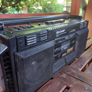 Può includere: Un registratore a cassette stereo Sharp nero con un doppio deck per cassette. Il registratore ha una piastra frontale argentata con il testo "SHARP" e "STEREO RADIO CASSETTE RECORDER" in bianco. Il registratore ha una griglia di altoparlanti in rete nera sul davanti.
