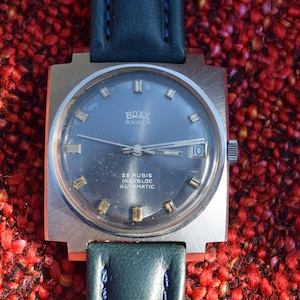 May include: A silver square-shaped wristwatch with a black face and a green leather band. The watch face has gold-colored hour markers and a date window at the 3 o'clock position. The watch face also has the text "ROXY ANKER 25 RUBIS INCABLOC AUTOMATIC".