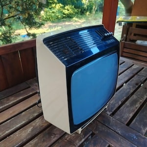 Pode incluir: Uma televisão antiga com corpo creme e parte superior preta. O ecrã é de cor azul clara. A TV está sobre uma superfície de madeira, com um fundo natural de árvores e folhagem.