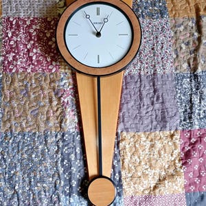 May include: A wooden Seiko wall clock with a light-coloured face, black hands, and a brown frame. The clock has a pendulum design with a black rod and a circular weight. The clock is mounted on a light-coloured wooden base.