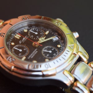 May include: A silver and gold chronograph wristwatch with a black face and a silver band. The watch face has three subdials and the text "Kolb Geneve Chronograph 1000 Meters" printed on it.