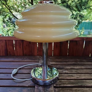 May include: A vintage table lamp with a chrome base and stem, topped with a tiered, cream-colored glass shade. The lamp is plugged in and sits on a wooden table, with a natural background of trees and foliage.
