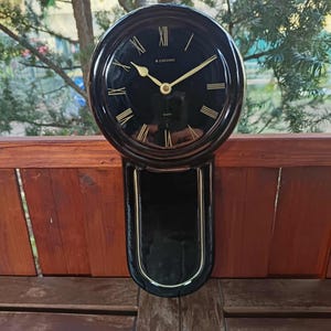 May include: A black quartz wall clock with gold-colored hands and Roman numerals. The clock has a round face and a rectangular pendulum. The brand name "Junghans" is visible on the clock face.