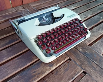 May include: A white vintage typewriter with a red and white keyboard. The typewriter is sitting on a wooden table.