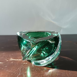 Può includere: Un posacenere decorativo in vetro verde smeraldo con un design unico e attorcigliato. Il posacenere ha una forma quadrata con bordi arrotondati e una ciotola profonda. Il vetro è trasparente, permettendo alla luce di passare e riflettersi.