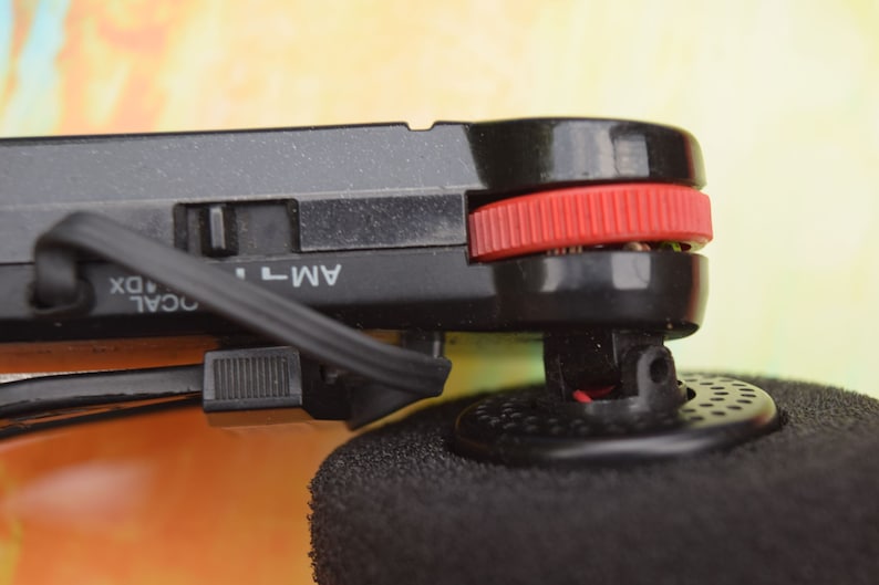 Pode incluir: Close-up de um r&aacute;dio preto com um bot&atilde;o vermelho e um cabo preto. O r&aacute;dio tem um r&oacute;tulo que diz "AM-T" e "LOCAL DX".