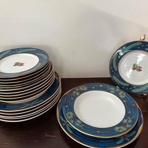 Pode incluir: Um conjunto de pratos azuis e brancos com borda dourada e design festivo. Os pratos têm uma borda azul escura com estrelas douradas e detalhes verdes e vermelhos. Uma pilha de pratos é visível, junto com dois pratos individuais.