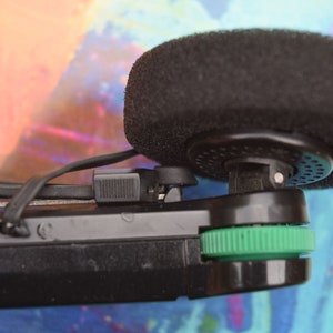 Pode incluir: Um microfone preto com um protetor de espuma preso a um dispositivo preto e verde.