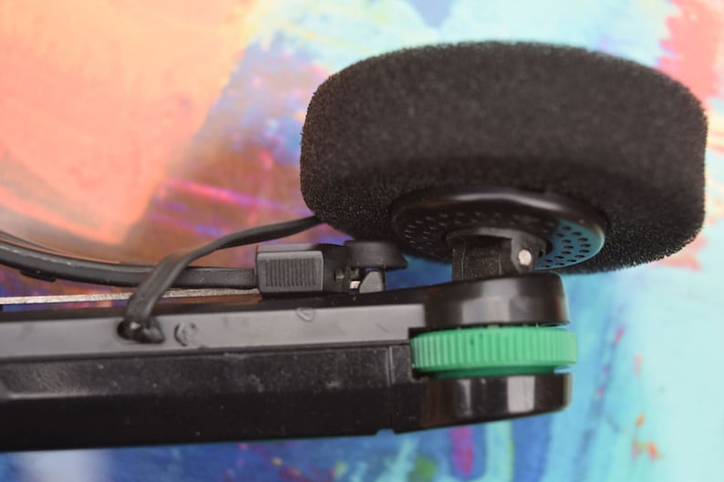 Pode incluir: Um microfone preto com um protetor de espuma preso a um dispositivo preto e verde.