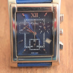 May include: A silver and blue chronograph wristwatch with a square face. The watch face has Roman numeral hour markers and a date window at the 4 o'clock position. The watch band is blue leather.
