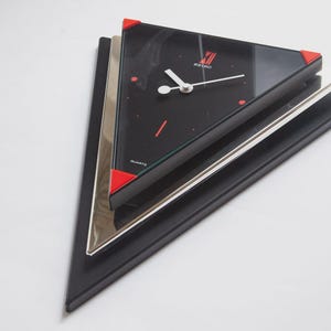 May include: A black and silver quartz clock with a triangular design. The clock face is black with white hands and red accents. The corners of the clock are red. The clock has the word "SEIKO" on the face.