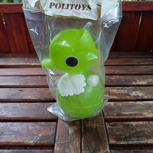 Puede incluir: Un juguete de plástico verde en una bolsa de plástico transparente. El juguete tiene forma de personaje de dibujos animados con una cabeza grande, ojos negros y un babero blanco. La bolsa lleva la etiqueta "POLITOYS" en letras marrones.