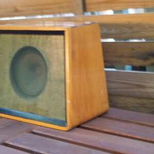 K&ouml;nnte beinhalten: Ein Vintage-Lautsprechergeh&auml;use aus Holz mit hellbraunem Finish. Die Vorderseite verf&uuml;gt &uuml;ber einen schwarz gerahmten Lautsprechergrill mit einem kreisf&ouml;rmigen Lautsprecher, der hinter dem Gitter sichtbar ist. Das Geh&auml;use ist abgewinkelt und steht auf einer Holzoberfl&auml;che.