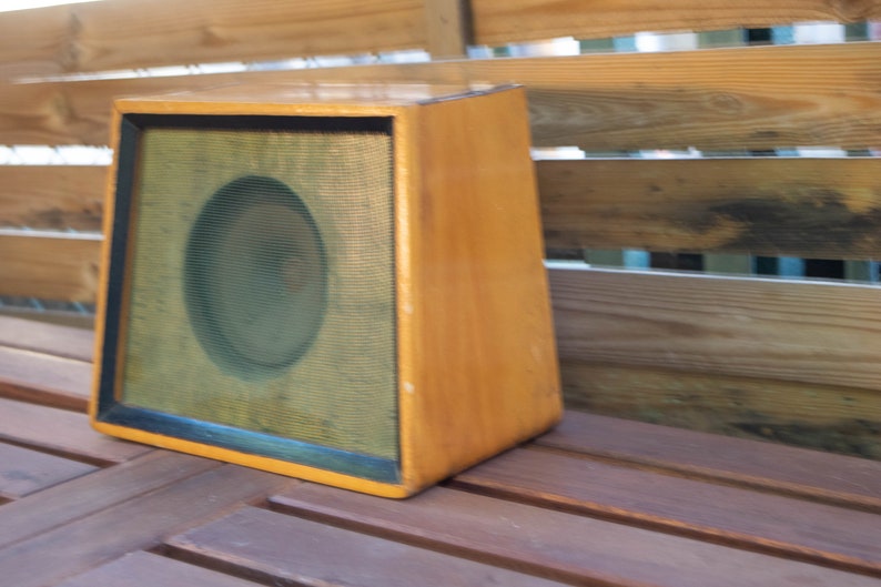 K&ouml;nnte beinhalten: Ein Vintage-Lautsprechergeh&auml;use aus Holz mit hellbraunem Finish. Die Vorderseite verf&uuml;gt &uuml;ber einen schwarz gerahmten Lautsprechergrill mit einem kreisf&ouml;rmigen Lautsprecher, der hinter dem Gitter sichtbar ist. Das Geh&auml;use ist abgewinkelt und steht auf einer Holzoberfl&auml;che.
