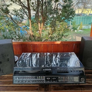 May include: A vintage black stereo system with two matching speakers. The system includes a turntable, radio, and cassette player. The speakers and stereo are on a wooden table, with a natural outdoor background.