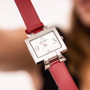 May include: A silver square watch with a white face and black hands. The watch has a red leather band.