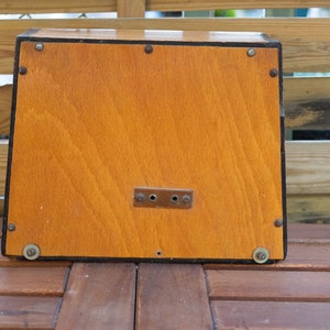 K&ouml;nnte beinhalten: Eine rechteckige Holzkiste mit hellbraunem Finish. Die Kiste hat einen dunklen Rand und ist mit kleinen Metallnieten befestigt. Eine kleine Metallplatte mit zwei L&ouml;chern ist vorne angebracht. Die Kiste steht auf einer Holzoberfl&auml;che.