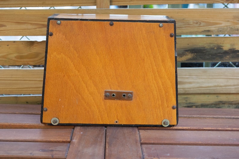 K&ouml;nnte beinhalten: Eine rechteckige Holzkiste mit hellbraunem Finish. Die Kiste hat einen dunklen Rand und ist mit kleinen Metallnieten befestigt. Eine kleine Metallplatte mit zwei L&ouml;chern ist vorne angebracht. Die Kiste steht auf einer Holzoberfl&auml;che.