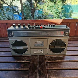 Könnte beinhalten: Ein silberner Vintage-Boombox mit schwarzen Akzenten, mit zwei großen Lautsprechern und einem Kassettenrekorder. Die Boombox hat mehrere Knöpfe und Tasten, und der Markenname "VIDEOTON" ist sichtbar. Sie steht auf einer Holzoberfläche.