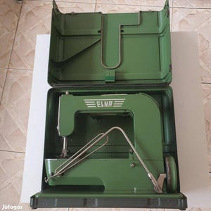 May include: A vintage green Elna sewing machine housed in its matching green carrying case. The machine features a prominent Elna logo on the side, a metal needle bar, and a metal presser foot lever. The case is open, revealing the machine inside.