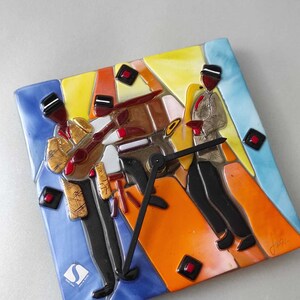 Könnte beinhalten: Quadratische Keramik-Wanduhr mit einer Jazzband-Illustration. Das Zifferblatt zeigt Musiker, die Instrumente spielen, mit einem farbenfrohen, abstrakten Design. Die Uhr hat schwarze Zeiger und einen blauen, orangefarbenen und gelben Hintergrund.