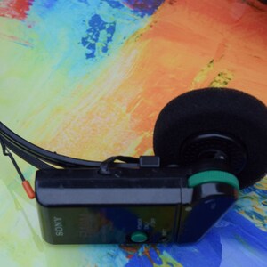 Pode incluir: Um est&eacute;reo pessoal Sony preto com um bot&atilde;o verde e um fone de ouvido de espuma preta. O est&eacute;reo est&aacute; em um fundo colorido.