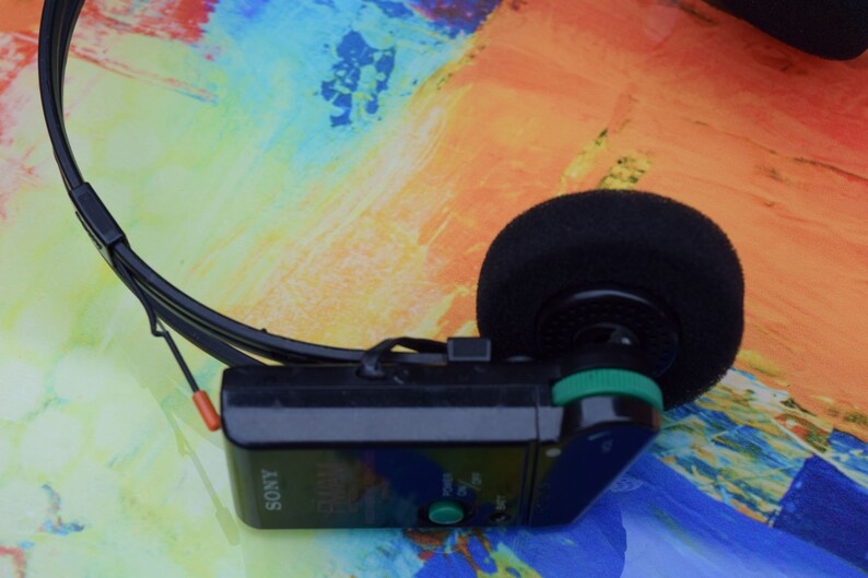 Pode incluir: Um est&eacute;reo pessoal Sony preto com um bot&atilde;o verde e um fone de ouvido de espuma preta. O est&eacute;reo est&aacute; em um fundo colorido.