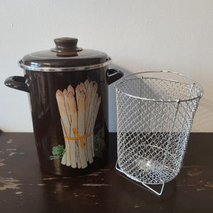 以下が含まれることがあります： 蓋と取っ手が付いた濃い茶色のエナメル鍋。黄色いリボンで束ねられたアスパラガスの絵が描かれています。その隣には、銀色のワイヤーメッシュバスケットがあります。