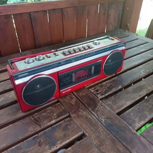 May include: A vintage red and silver Sharp Q12 boombox with two black speakers and a cassette player. The boombox has a silver control panel with various buttons and knobs. The boombox is sitting on a wooden table.