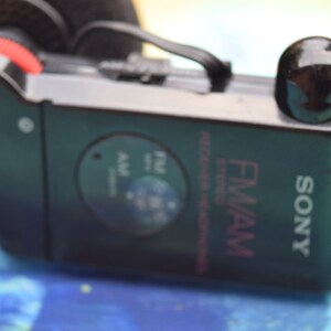 Pode incluir: Um r&aacute;dio est&eacute;reo de bolso FM/AM Sony preto com uma tomada de auscultadores preta e um bot&atilde;o preto para sintonizar. O r&aacute;dio tem um r&oacute;tulo prateado com o texto "SONY" e "FM/AM" em rosa.