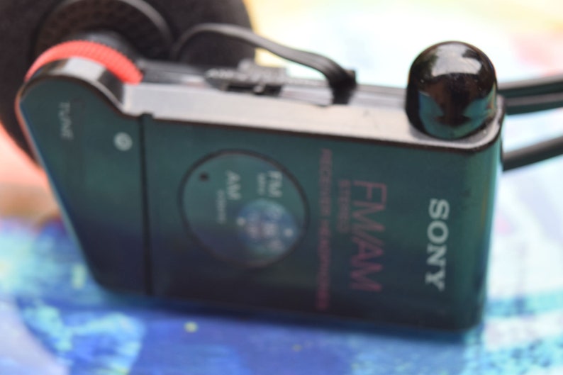 Pode incluir: Um r&aacute;dio est&eacute;reo de bolso FM/AM Sony preto com uma tomada de auscultadores preta e um bot&atilde;o preto para sintonizar. O r&aacute;dio tem um r&oacute;tulo prateado com o texto "SONY" e "FM/AM" em rosa.