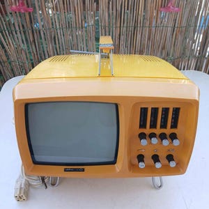 以下が含まれることがあります： 鮮やかな黄色のポータブルテレビ。小さな画面、上部の持ち運び用ハンドル、右側のコントロールパネルには4つの垂直チャンネルスライダーと6つの丸いノブが付いています。画面の下にはMINI-VIDというブランド名が印刷されています。