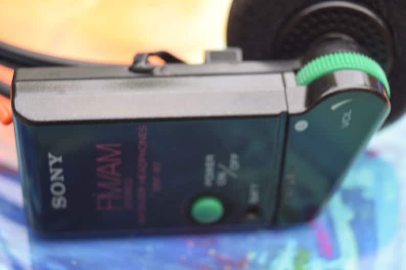 Pode incluir: Um fone de ouvido est&eacute;reo FM/AM Sony preto com um bot&atilde;o de volume verde e um bot&atilde;o de energia verde. Os fones de ouvido t&ecirc;m um r&oacute;tulo rosa e branco que diz "FM/AM Stereo Headphones".