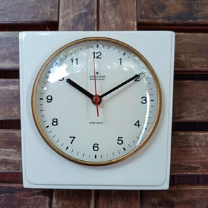 Peut inclure: Une horloge murale carrée blanche avec une lunette dorée et des aiguilles noires. Le cadran de l'horloge comporte des chiffres noirs et la marque "JUNGHANS electronic" est visible. L'horloge indique "ATO-MAT".