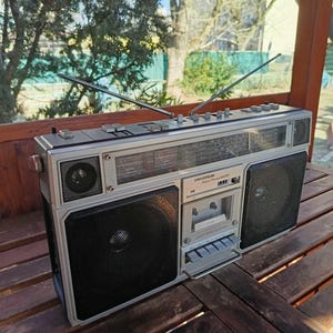 Puede incluir: Un boombox vintage plateado con un reproductor de casetes y un sintonizador de radio. El boombox tiene dos altavoces y un panel de control plateado con botones y perillas. El boombox está sentado sobre una superficie de madera.