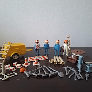 Op de afbeelding: Een Playmobil constructiespeelgoed set met figuren en accessoires. Inclusief een gele aanhanger, ladders, verkeerskegels en diverse gereedschappen. De figuren dragen blauwe en witte uniformen, ideaal voor fantasierijk spel.