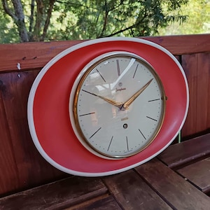 May include: A vintage wall clock with a red oval frame and a white border. The clock face is cream-colored with black hour markers and gold hands. The clock face has the word "Junghans" printed on it.