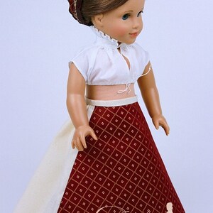 May include: A doll wearing a white crop top with a ruffled collar and a red patterned skirt. The skirt has a diamond pattern with gold accents. A matching red headband completes the outfit. The doll is a classic toy.