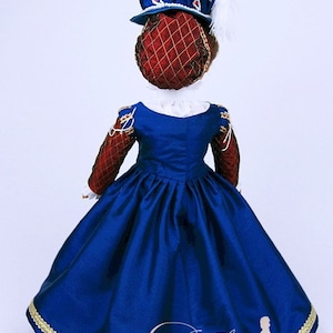 May include: A doll wearing a historical costume, featuring a royal blue dress with a full skirt and gold trim. The bodice has red and gold sleeves. A matching hat with a red net and blue trim is on the doll's head. A white feather is attached to the hat.