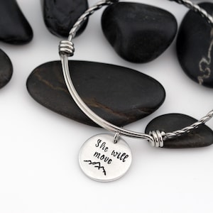 May include: A silver bangle bracelet with a charm that reads "She will move" and a small mountain range graphic.