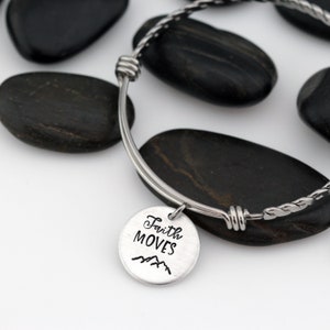 May include: A silver bangle bracelet with a silver charm that says "Faith Moves" with a mountain range design.