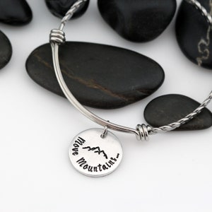 May include: A silver bangle bracelet with a charm that reads "I have mountains..." with a mountain silhouette graphic. The bracelet is on a white surface with black stones.