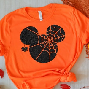 May include: Bright orange t-shirt featuring a black Mickey Mouse head design with a spider web and a hanging spider. The shirt is styled with rolled-up sleeves and a knot at the bottom. Pumpkins and fall foliage are arranged around the shirt.