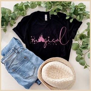 Peut inclure: Un t-shirt noir avec un motif paillet&eacute; rose du ch&acirc;teau de Cendrillon et le mot "magical". Le t-shirt est pos&eacute; sur une surface blanche avec un jean bleu et un chapeau de paille.
