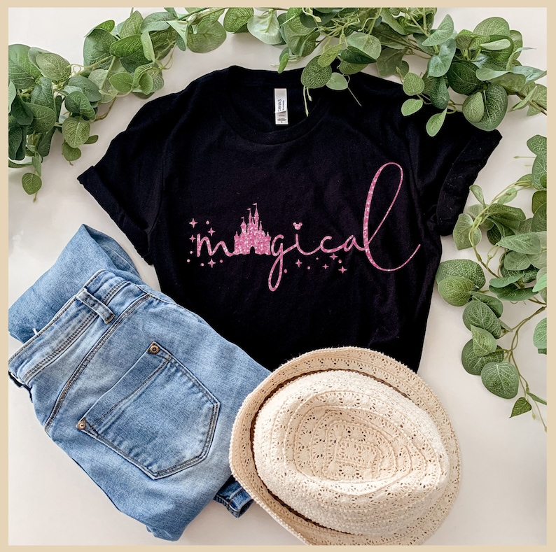 Peut inclure: Un t-shirt noir avec un motif paillet&eacute; rose du ch&acirc;teau de Cendrillon et le mot "magical". Le t-shirt est pos&eacute; sur une surface blanche avec un jean bleu et un chapeau de paille.