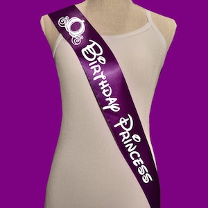 Puede incluir: Banda de sat&eacute;n morado con texto blanco que dice "Be Birthday Princess" y una silueta blanca de un carruaje y una corona.
