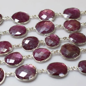 Könnte beinhalten: Eine Kette aus facettierten Rubin-Edelsteinen, die durch eine silberne Kette verbunden sind. Die Perlen sind oval geformt und haben eine tiefrote Farbe.
